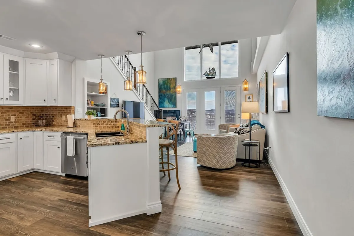 A spacious kitchen and gathering area anchored by a large central island, surrounded by comfortable seating and illuminated by natural light streaming through large windows. The space invites both socializing and meal prep, with decorative art accents and a cozy ambiance ideal for group activities.