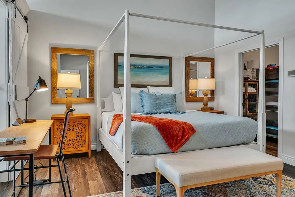 A welcoming king bedroom featuring a four-poster bed adorned with decorative pillows and a colorful throw blanket. The room includes decorative mirrors, a desk with a chair for personal or work use, and a table lamp, creating a serene environment for rest and productivity.