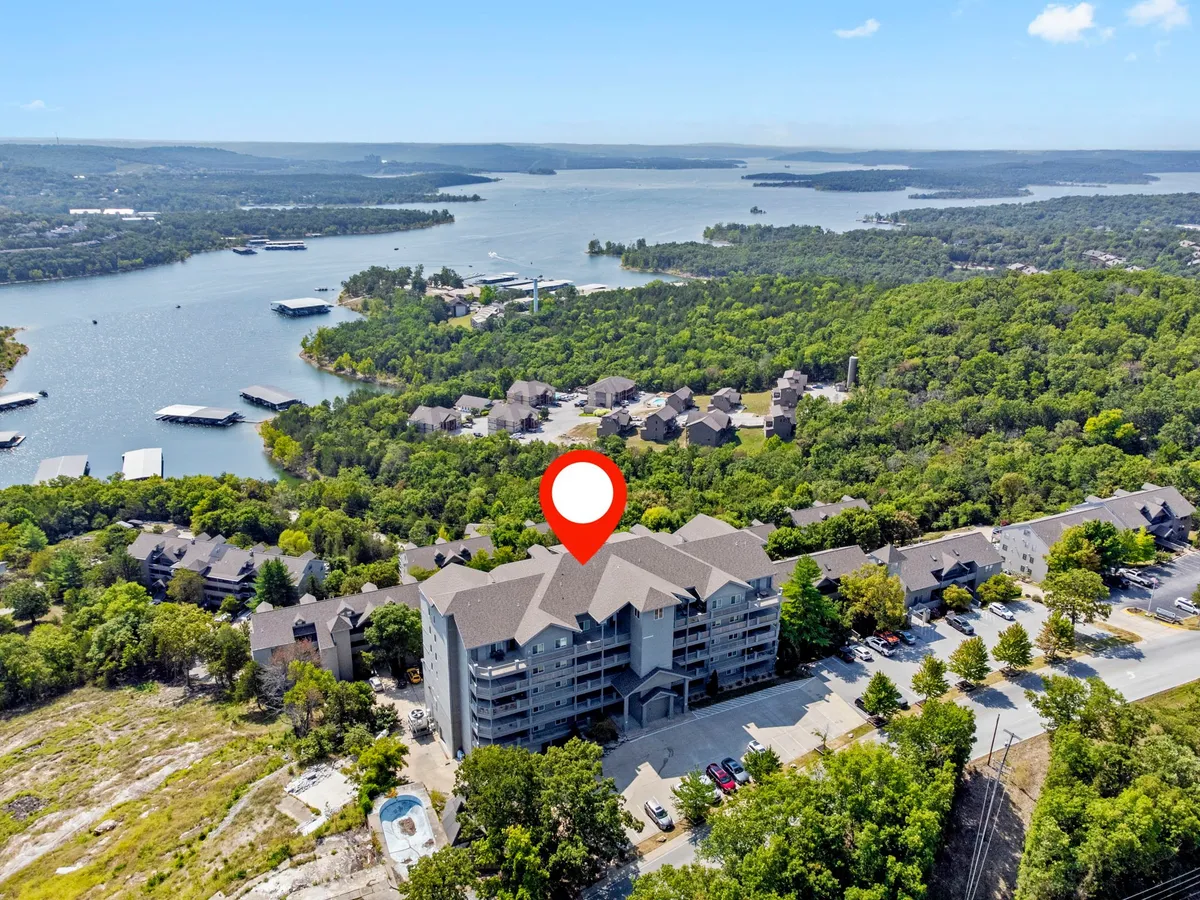 A tranquil exterior scene featuring Table Rock Lake bordered by a wooded area, with residential buildings and a parking lot visible. The calm water reflects the sky and surrounding trees, evoking a sense of lakeside adventure and community.
