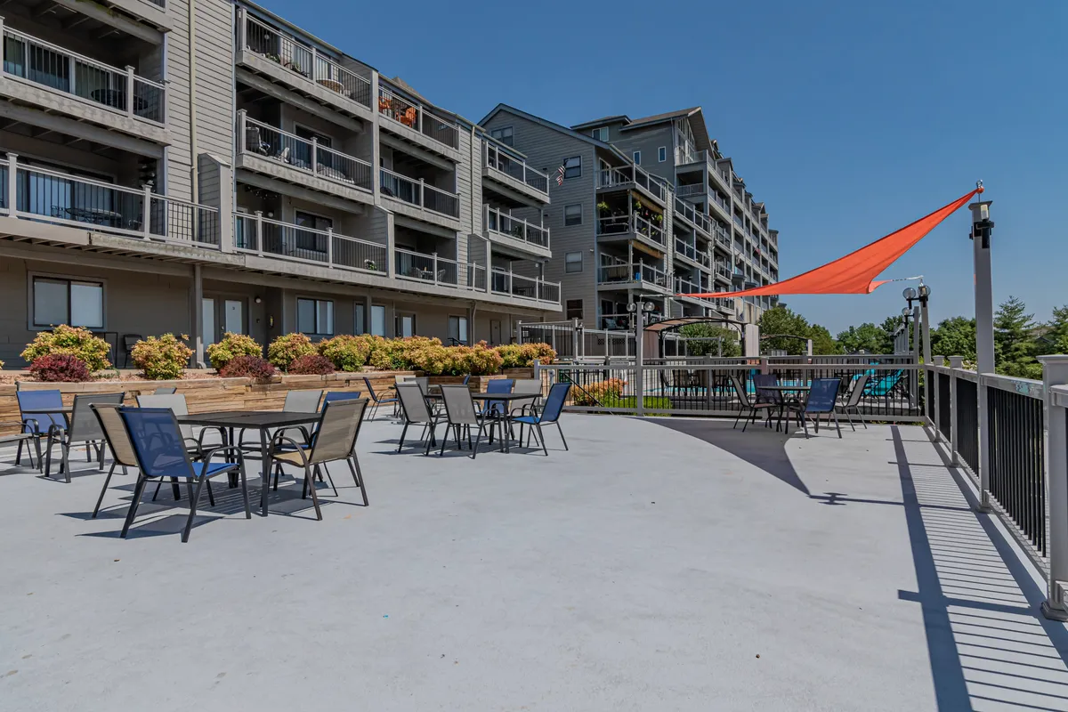 A lively outdoor seating area adorned with an orange shade sail, vibrant flower planters, and balconies in the background. The scene is set for social gatherings, outdoor dining, and relaxed sun-filled moments.