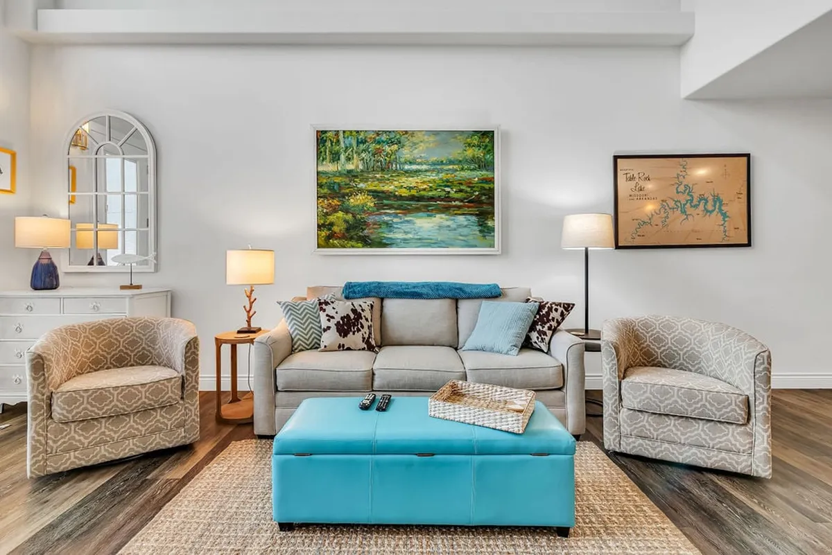The image showcases a stylish, inviting living area with a plush sofa and blue ottoman, accented by elegant artwork and a warm lamp. The space is filled with natural light, creating a cozy atmosphere that's perfect for relaxing, socializing, or unwinding with a good book. The updated finishes and thoughtful decor hint at a modern yet comfortable retreat.