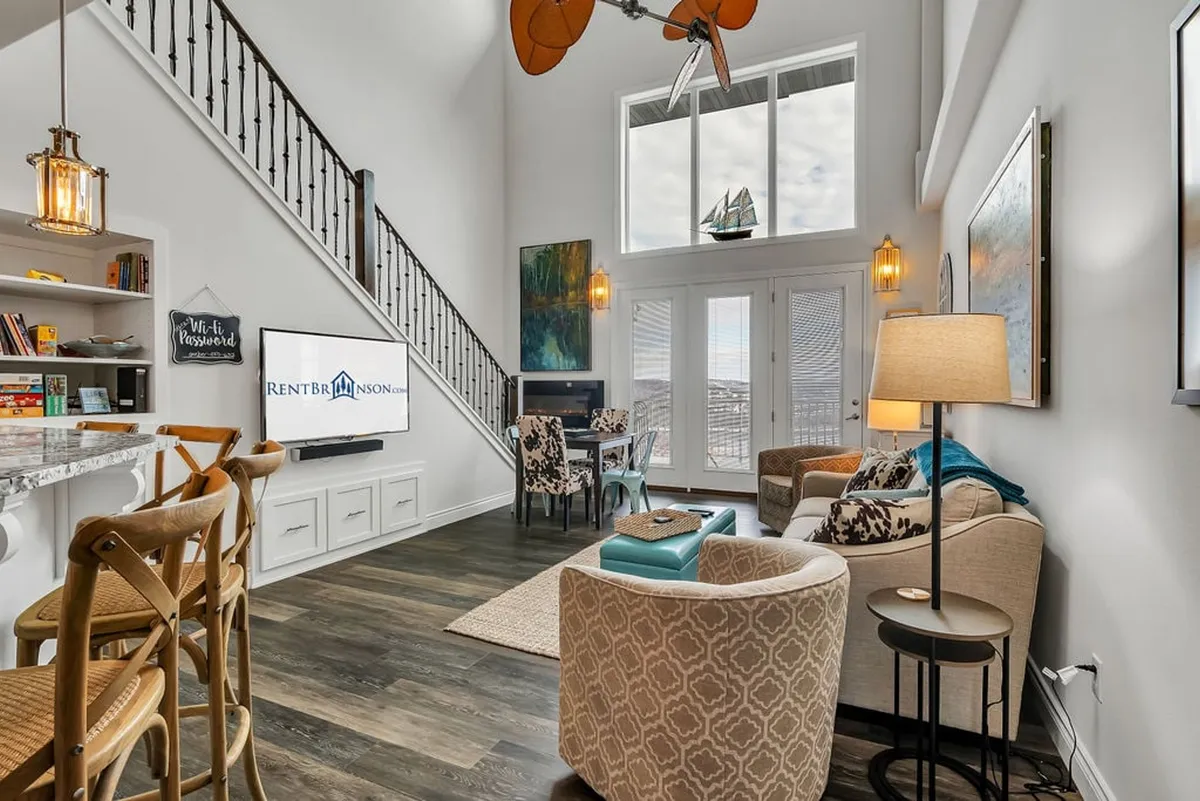 A comfortable living room filled with natural light from large windows, featuring cozy seating, a stylish ceiling fan, decorative lighting, and modern art accents. The space is designed for relaxation, socializing, and enjoying the view.