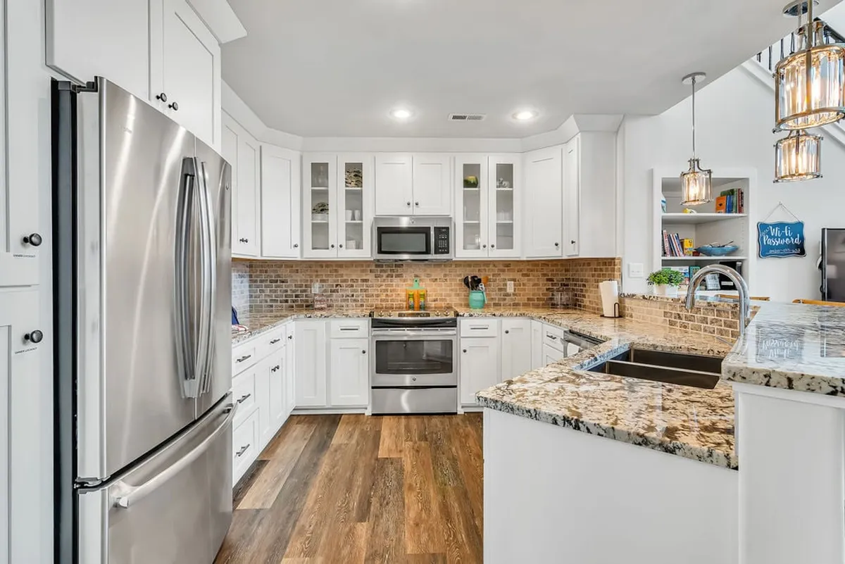 This image features a modern kitchen with gleaming stainless steel appliances, expansive granite countertops, crisp white cabinetry, and stylish decorative lighting fixtures. The inviting space is designed for cooking, entertaining, and family gatherings, offering a welcoming atmosphere at the very heart of the home.
