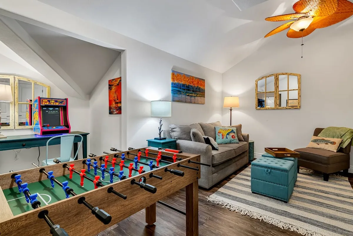 This image captures a vibrant and fun game room with a foosball table, an arcade game machine, a cozy seating area, decorative artwork, and a unique ceiling fan. The environment is bright, welcoming, and clearly designed for socializing and entertainment.
